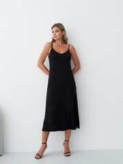 Woman in a black sleeveless dress with spaghetti straps, standing with hands behind her back; wearing black sandals. She gazes to the side with minimal, natural makeup, long earrings