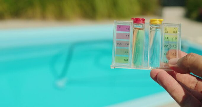 Close-up of a hand holding a water test kit for pH and chlorine levels against a clear blue swimming pool background, emphasizing pool maintenance.