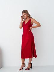 Woman in elegant red dress poses with hand on hip, looking down. She wears black high heels and a stylish necklace. Her long hair cascades over shoulders, exuding sophistication