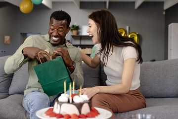 Opening gift bag, African American man and woman laughing, celebrating together on couch