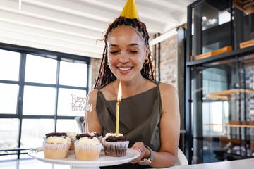 Mid-adult woman holding cupcakes with lit candle and glittery sign in loft, celebrating birthday