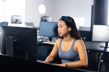 Asian woman working on large computer monitor at desk in open-plan office, with keyboard and mouse