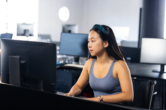 Asian woman working on large computer monitor at desk in open-plan office, with keyboard and mouse
