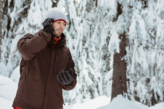 Fototapeta man talking on smartphone in snowy evergreen forest, wearing knit beanie and insulated jacket