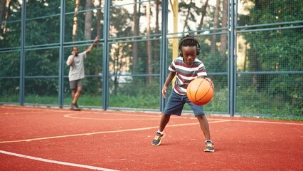 Little African American cute kid boy playing basketball outdoor on court. Small child listening to music in headphones while play basketball on street. Father talking on smartphone on background