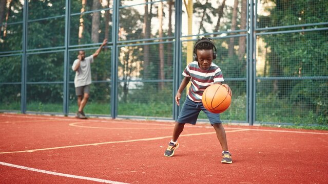 Little African American cute kid boy playing basketball outdoor on court. Small child listening to music in headphones while play basketball on street. Father talking on smartphone on background