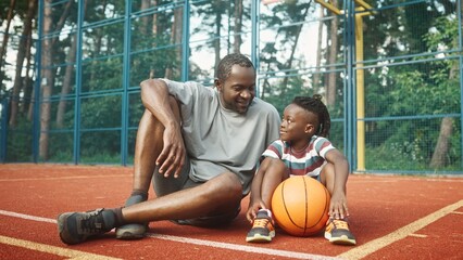 Happy little boy sitting outdoor on basketball court with father looking at camera and smiling. Family time. Dad and son with ball played basketball outside. Sport and recreation. Leisure concept