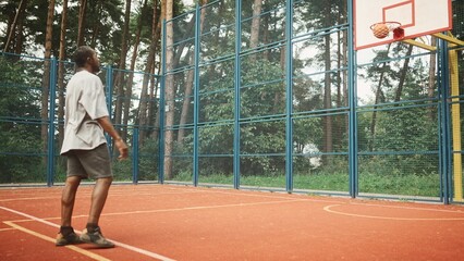 Mature adult African American sporty male practicing basketball throwing ball into hoop. Rear of professional player playing basketball hitting the hoop. Outdoor court. Urban playground in park