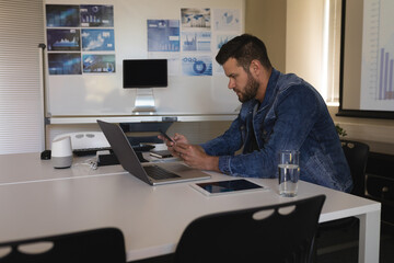 Mid adult man checking smartphone and laptop in meeting room, with projection screen, copy space