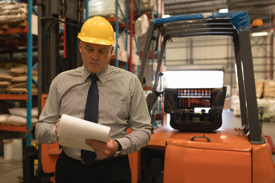 Senior male warehouse supervisor reviewing paperwork in aisle, with forklift truck, copy space
