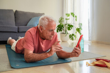 Elderly man lying down on yoga mat following workout instructions on his smartphone at home