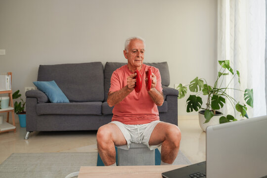 Senior man exercising with dumbbells at home watching online training on laptop