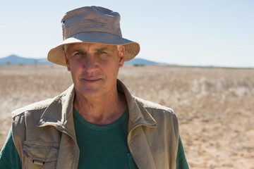 Standing senior man wearing green t-shirt and utility vest in arid plain, with bucket hat