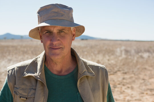 Standing senior man wearing green t-shirt and utility vest in arid plain, with bucket hat