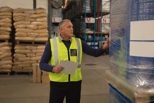Senior man examining wrapped pallet in warehouse, with clipboard and burlap sacks