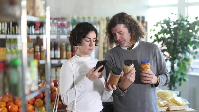 Spouses gets acquainted with food scan QR code, take reading canned beans and lentils label using phone camera in shop. Client view legume package, inspect manufacturer information using mobile app