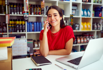 Young saleswoman smiling and talking on the phone while working at the counter of her auto parts...