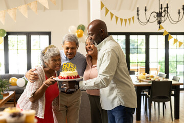 Holding cake, diverse family celebrating 60th birthday and smiling in decorated room