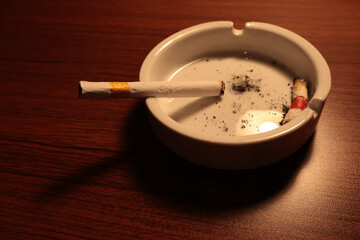 A cigarette rests in a white ashtray, surrounded by ash and the remnants of a finished smoke. The dim lighting adds a moody, contemplative atmosphere.