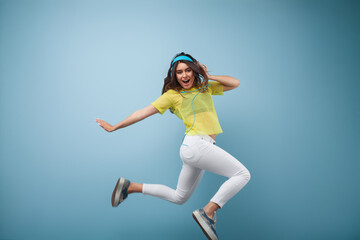 Fototapeta premium A vibrant young woman wearing headphones and energetically jumping against a blue background, exuding fun and carefree vibes.