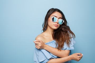A fashionable woman wearing sunglasses and a stylish off-shoulder blouse, standing confidently against a soft pastel blue backdrop, embodying modern style and sophistication.