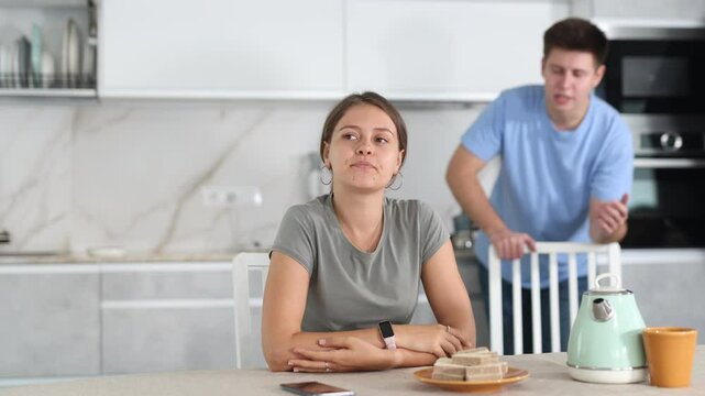 Sad young girl sitting at the table while her boyfriend is quarreling with her standing behind in the kitchen