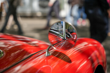 Fototapeta premium A close-up of a red vintage car's round chrome mirror. The mirror reflects its windshield and chrome trim, capturing the movement of people in the background.