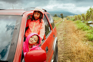Two Asian little girl peek out from car window and smiling, enjoy their road trip adventure to the mountain track