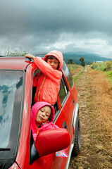 Two little Asian sisters peeking out from car window and smiling, enjoying their adventurous road trip to through the mountain track
