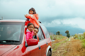 Happy Asian mother with her two little daughters peeking out from car window, enjoying nature scenery on their road trip adventure through the mountain track
