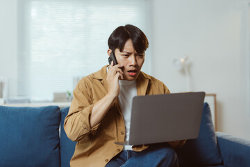 Young man experiencing frustration during a phone call while working from home, sitting on the sofa and focused on his laptop