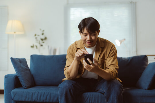 Happy asian man using mobile phone browsing internet, ordering online shopping, checking social media, playing game, chatting with friends at home