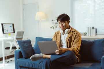 Young asian man working from home sitting on the sofa and using laptop, wearing headphones and...