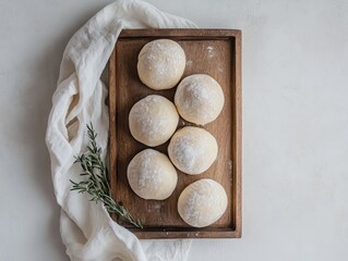 Six round, pale dough balls arranged on a wooden tray, dusted with powder, beside a sprig of herbs and linen cloth
