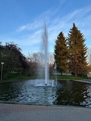 Fototapeta premium Vertical photo of a fountain in a peaceful city park in Warsaw, Poland. Surrounded by coniferous and deciduous trees under a clear blue sky in the evening light