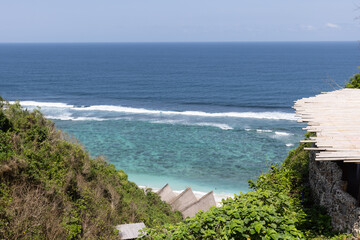 Wide high angle shot of beach club in Bali with straw roofs next to the ocean with rolling waves on a sunny day with blue skies in Uluwatu, Bali, Indonesia. 