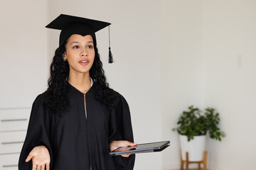 Graduate in cap and gown holding tablet, speaking confidently at home