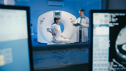 Dark-haired female sitting upright near CT scanner and resting hands on lap while awaiting feedback. Thoughtful physician holding chin and reviewing diagnostic notes behind glass with scans displayed. © ihorvsn