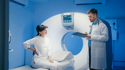 Serious Caucasian female sitting on CT scanner and listening carefully to focused male doctor...
