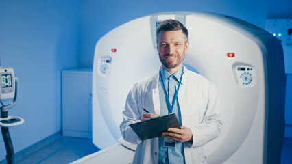 Positive Caucasian male in medical coat smiling and holding clipboard while posing near CT scanner. Preparing for next appointment or reviewing patient files inside clinical diagnostic center.