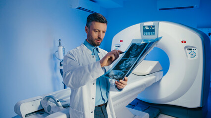 Skilled doctor holding pen and pointing at brain scan film during image evaluation. Handsome Caucasian male reviewing diagnostic results next to CT machine. Concentrating inside radiology room.