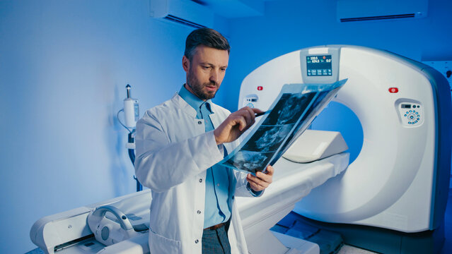 Skilled doctor holding pen and pointing at brain scan film during image evaluation. Handsome Caucasian male reviewing diagnostic results next to CT machine. Concentrating inside radiology room.