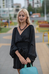 Fototapeta premium Portrait of a young beautiful blonde girl in a dark dress in a city dress in the summer.
