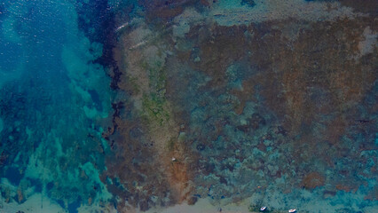 Aerial drone view of a vibrant blue sea with patches of coral reefs and seaweed visible beneath the clear, transparent water. Natural marine textures and patterns.