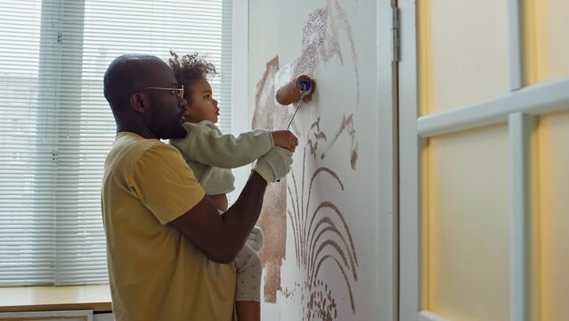 African American father holding young daughter while together painting walls using roller in home. Child eagerly helping, spreading paint on wall creating patterns