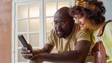 Couple smiling and looking at smartphone inside home with natural lighting coming through window. Man in casual t-shirt and woman in patterned headband both seem engaged with screen