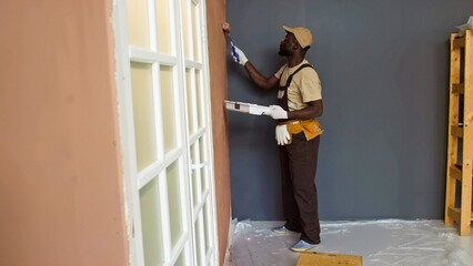 Black person skillfully painting house interior walls while standing on protective sheet. Walls...