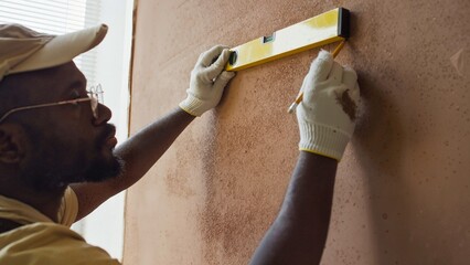 Man wearing gloves and cap carefully using a level tool for precise wall measurements in home...