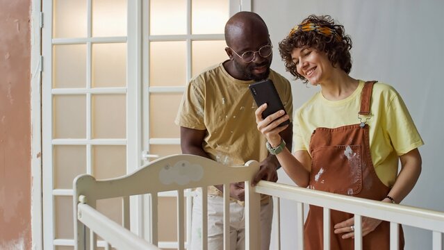 Expecting parents standing in nursery, looking at smartphone while working on baby crib. Father in yellow shirt and mother in orange overalls, both engaged in activity - Powered by Adobe