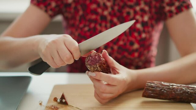 Professional chef hand scrapes cured meat casing to reveal tender sausage inside on wooden board, soft window light highlighting precise culinary technique and textured meat surface